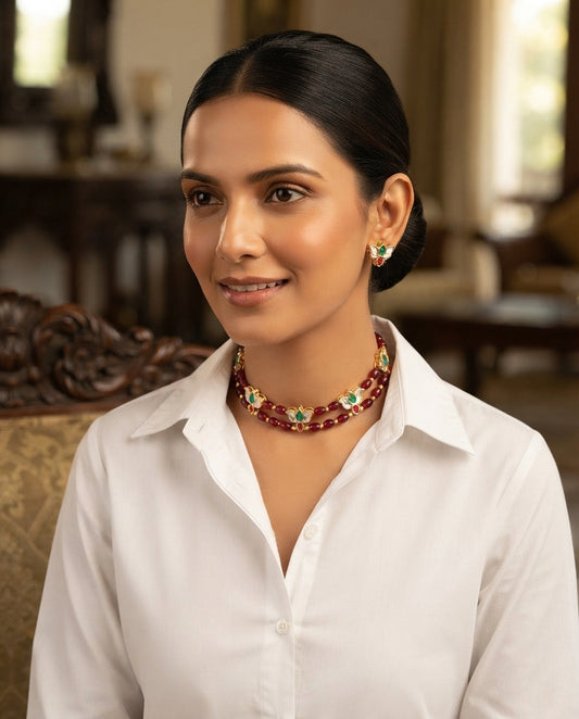 A woman with dark hair pulled back wears a white collared shirt and a red beaded necklace with gold and green decorative details, posed in an elegant indoor setting.
