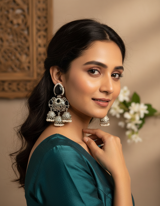 A woman wearing ornate silver-toned jhumka earrings with white stones and pearl-like dangling bells, photographed in a warm indoor setting with a carved wooden panel and white flowers in the background.
