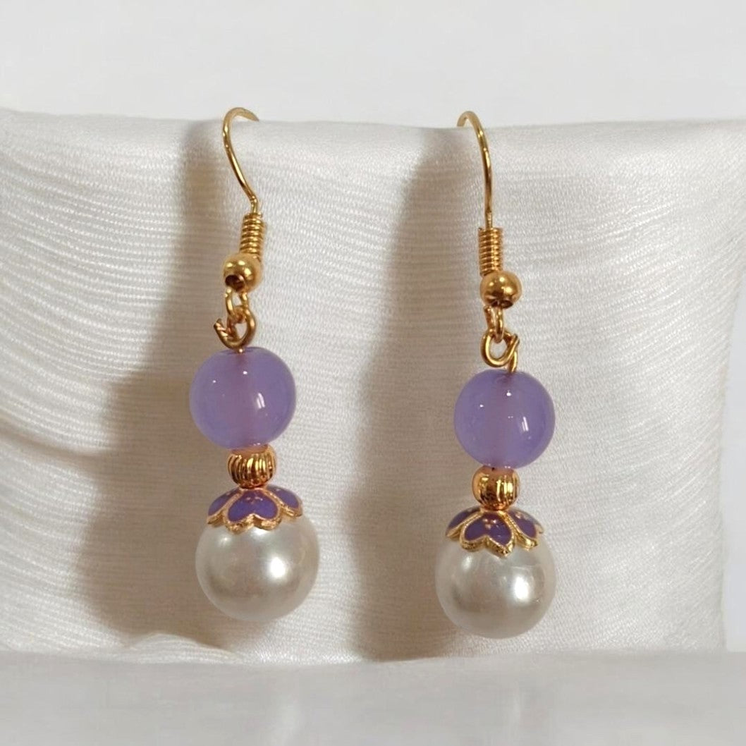 A pair of gold-tone dangle earrings with light purple beads, a small gold floral accent, and a white pearl drop, displayed on a white satin stand.