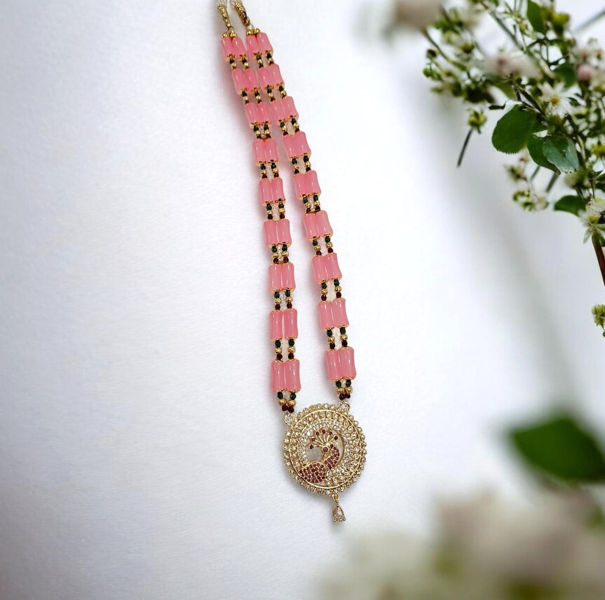 A pink and gold beaded necklace with a circular Om pendant, styled on a white surface with soft green and white flowers in the background.