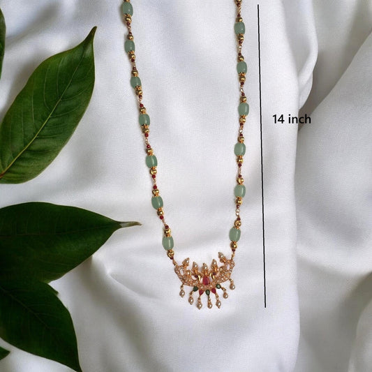 A gold-toned beaded necklace with light green oval beads and a decorative pendant, displayed on a white fabric background with green leaves. A vertical measurement line and text indicate the necklace is 14 inches long.