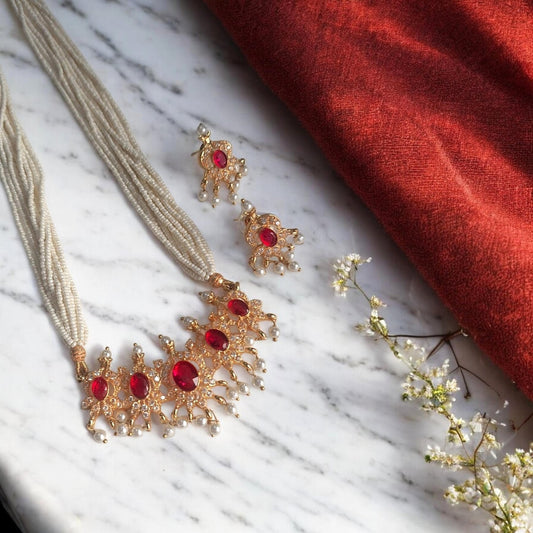 Gold-toned traditional jewelry set with a multi-strand necklace and matching earrings, each featuring red gemstone accents, arranged on a white marble surface next to a red fabric drape and small white flowers.