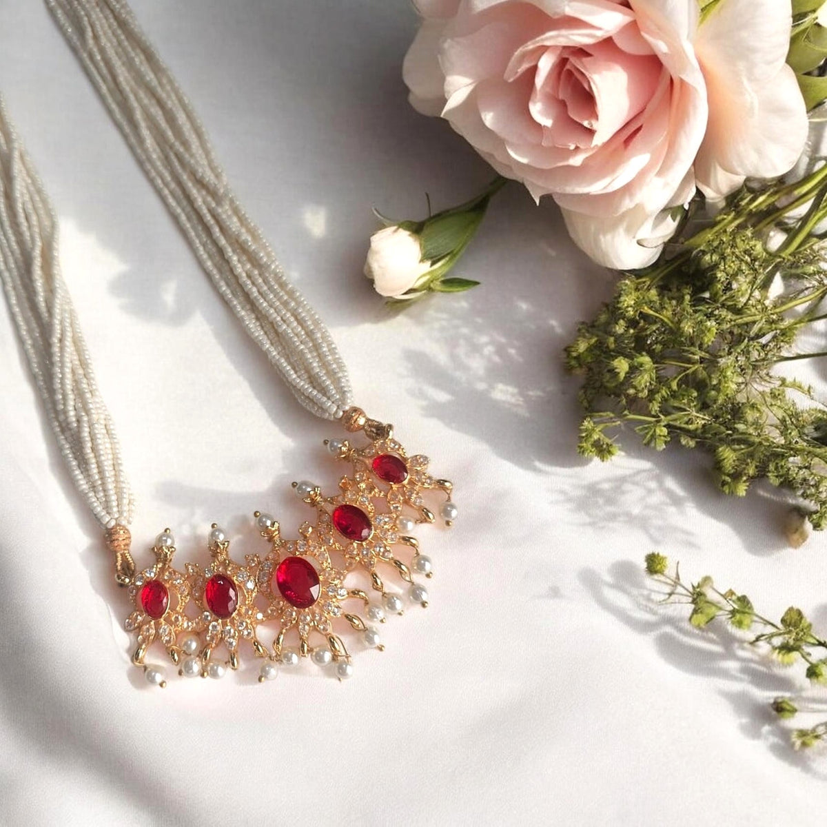A gold-toned statement necklace with multiple red oval gemstones and clear crystal-like accents, displayed on a white fabric surface with pink and white flowers as styling props.