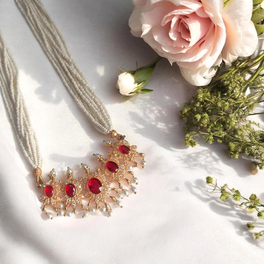A gold-toned statement necklace with multiple red oval gemstones and clear crystal-like accents, displayed on a white fabric surface with pink and white flowers as styling props.