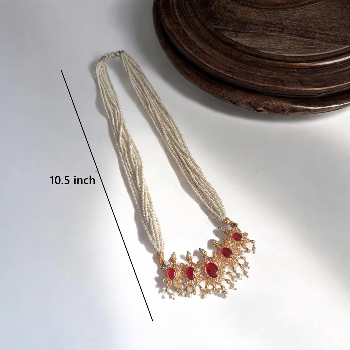 A multi-strand cream-colored beaded necklace with a gold-toned crescent pendant featuring red gemstones, shown on a white surface with a wooden tray in the background and a '10.5 inch' size overlay.