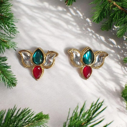 A pair of gold-toned butterfly-shaped earrings with a teal marquise stone, a red teardrop stone, and clear faceted side stones, arranged on a white textured surface with pine branches around the edges.