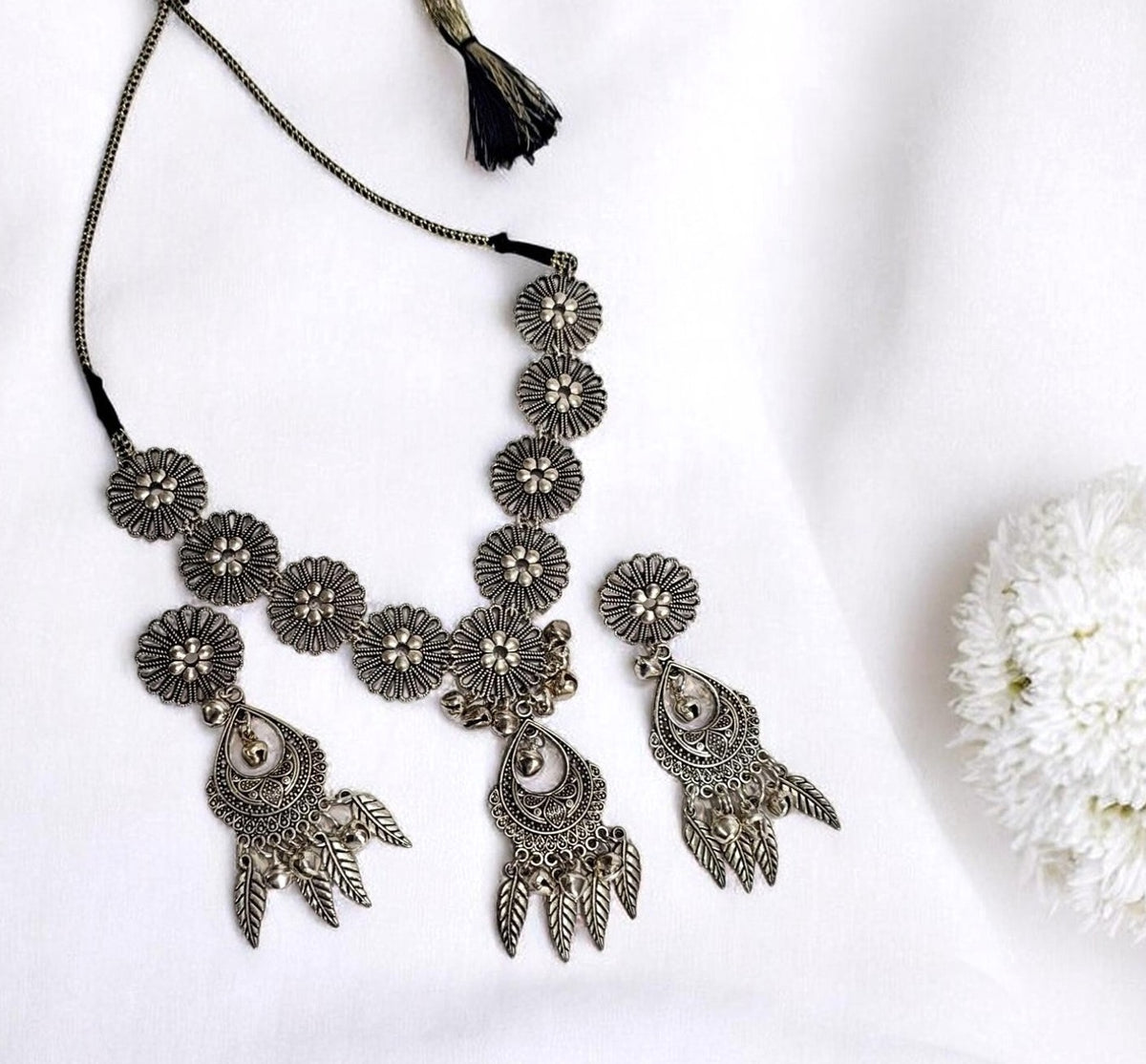 An ornate silver-toned bohemian-style necklace with matching earrings, featuring intricate filigree details, floral motifs, and dangling leaf-like charms, displayed on a white fabric background with a decorative white flower.