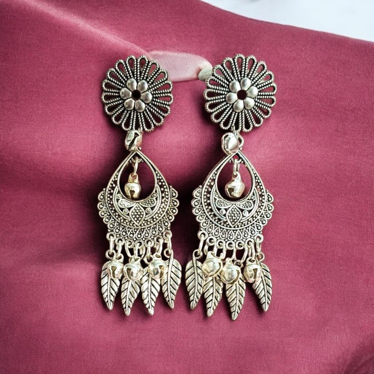 A pair of ornate silver-toned dangle earrings with floral studs, filigree teardrop pendants, and leaf-shaped dangling charms, displayed on a textured magenta fabric background.