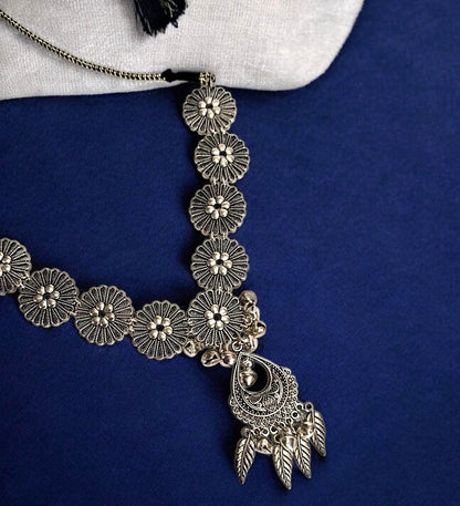 An antique silver-toned floral necklace with a silver oxidised pendant, displayed on a textured blue surface with a white fabric accent at the top.