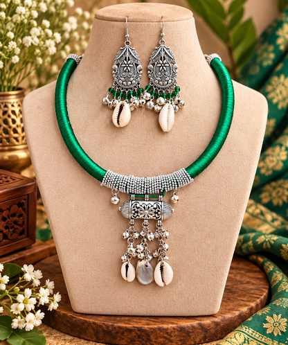 A traditional Indian jewelry set featuring a statement necklace and matching earrings displayed on a beige mannequin bust. The necklace has a vibrant green beaded collar, a silver-toned textured center, and dangling cowrie shells and silver filigree.