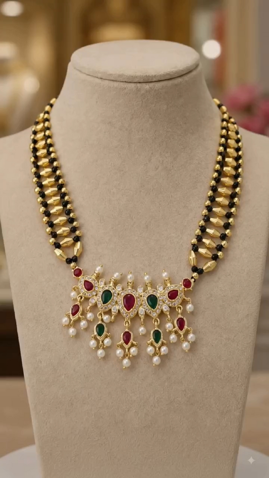 A gold-plated statement necklace displayed on a beige jewelry bust. The necklace features a multi-strand design with gold and black beads, a central gold collar decorated with red and green stones, and dangling pearl-like accents.