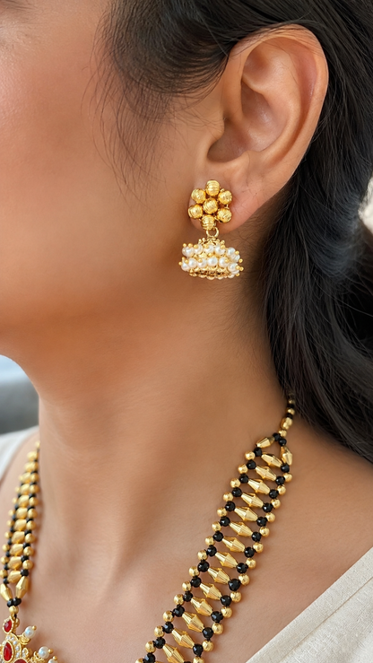 A close-up of a woman wearing a gold and white floral jhumka earring, paired with a matching gold and black beaded necklace.
