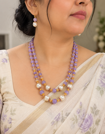 A close-up of a woman wearing a double-strand beaded necklace and matching drop earrings. The jewelry features lavender purple beads, white pearls, and small gold spacer beads.