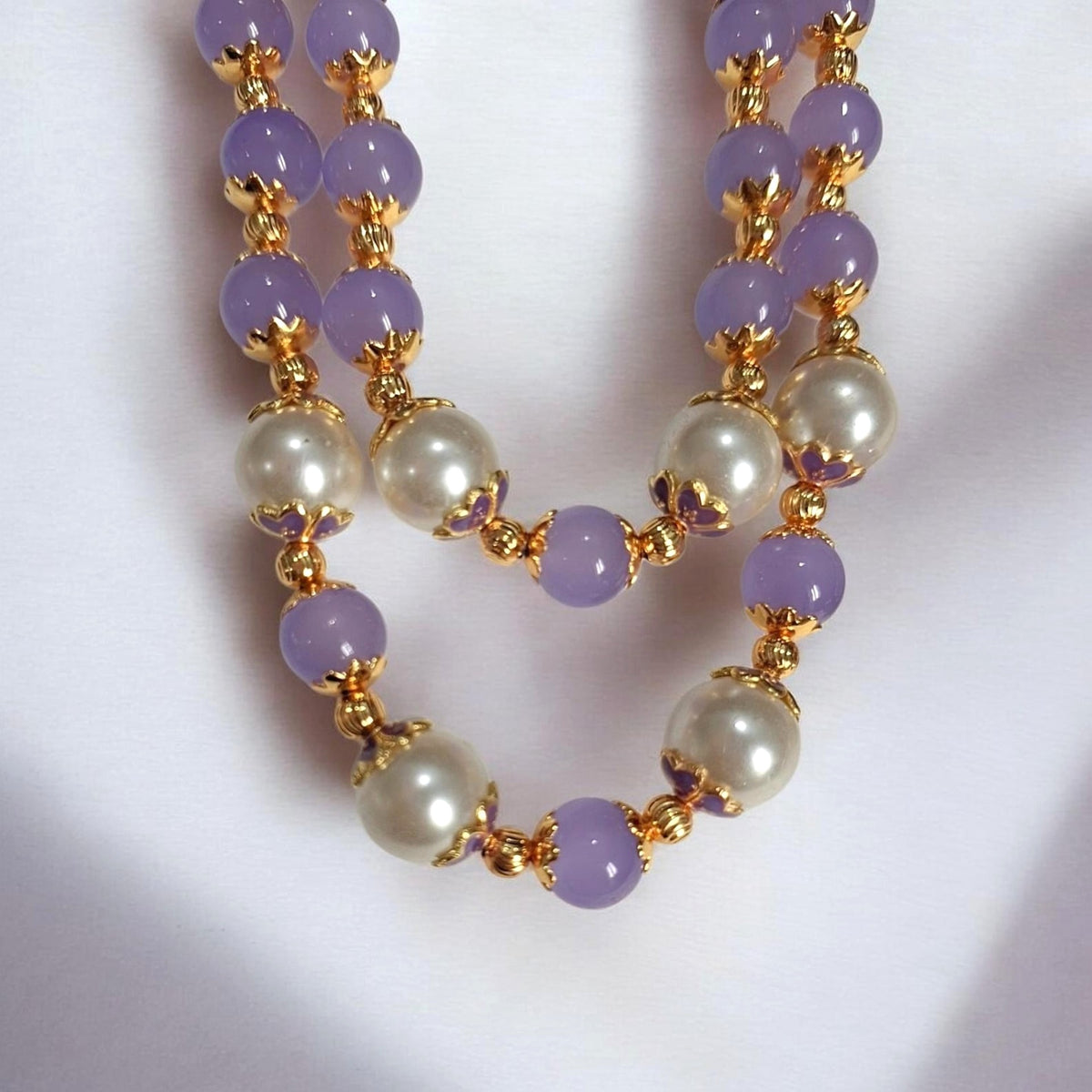 A double-strand beaded necklace featuring lavender purple beads, white faux pearls, and gold-toned spacer rings, displayed on a white fabric background.