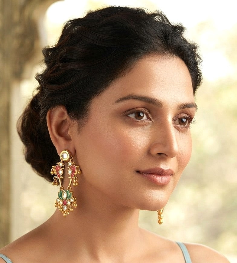 A woman wearing ornate gold-toned traditional Indian earrings with green, pink, and white stones, shown in a close-up portrait against a softly blurred outdoor background.