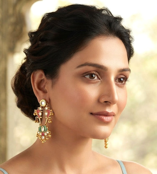 A woman wearing ornate gold-toned traditional Indian earrings with green, pink, and white stones, shown in a close-up portrait against a softly blurred outdoor background.