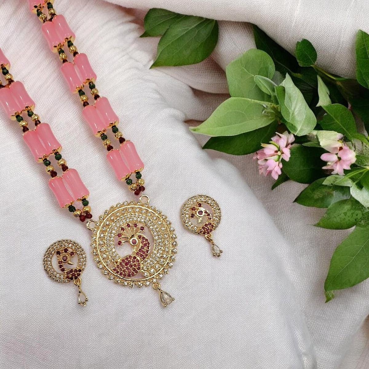 Gold-plated peacock necklace with pink stone beads and matching round earrings, arranged on a white fabric background with green leaves and small pink flowers.