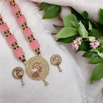 Gold-plated peacock necklace with pink stone beads and matching round earrings, arranged on a white fabric background with green leaves and small pink flowers.