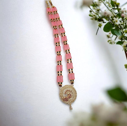 A pink and gold beaded necklace with a circular Om pendant, styled on a white surface with soft green and white flowers in the background.