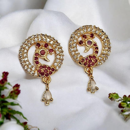 A pair of gold-toned Indian-style earrings with circular pavé crystal borders, red gemstone peacock motifs, and dangling clear teardrop crystals, displayed on a white fabric background with small floral accents.