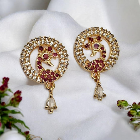 A pair of gold-toned Indian-style earrings with circular pavé crystal borders, red gemstone peacock motifs, and dangling clear teardrop crystals, displayed on a white fabric background with small floral accents.