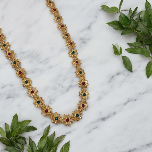 Sumana gold plated long necklace with ruby red and emerald green floral motifs, South Indian bridal jewellery, flat lay on white background
