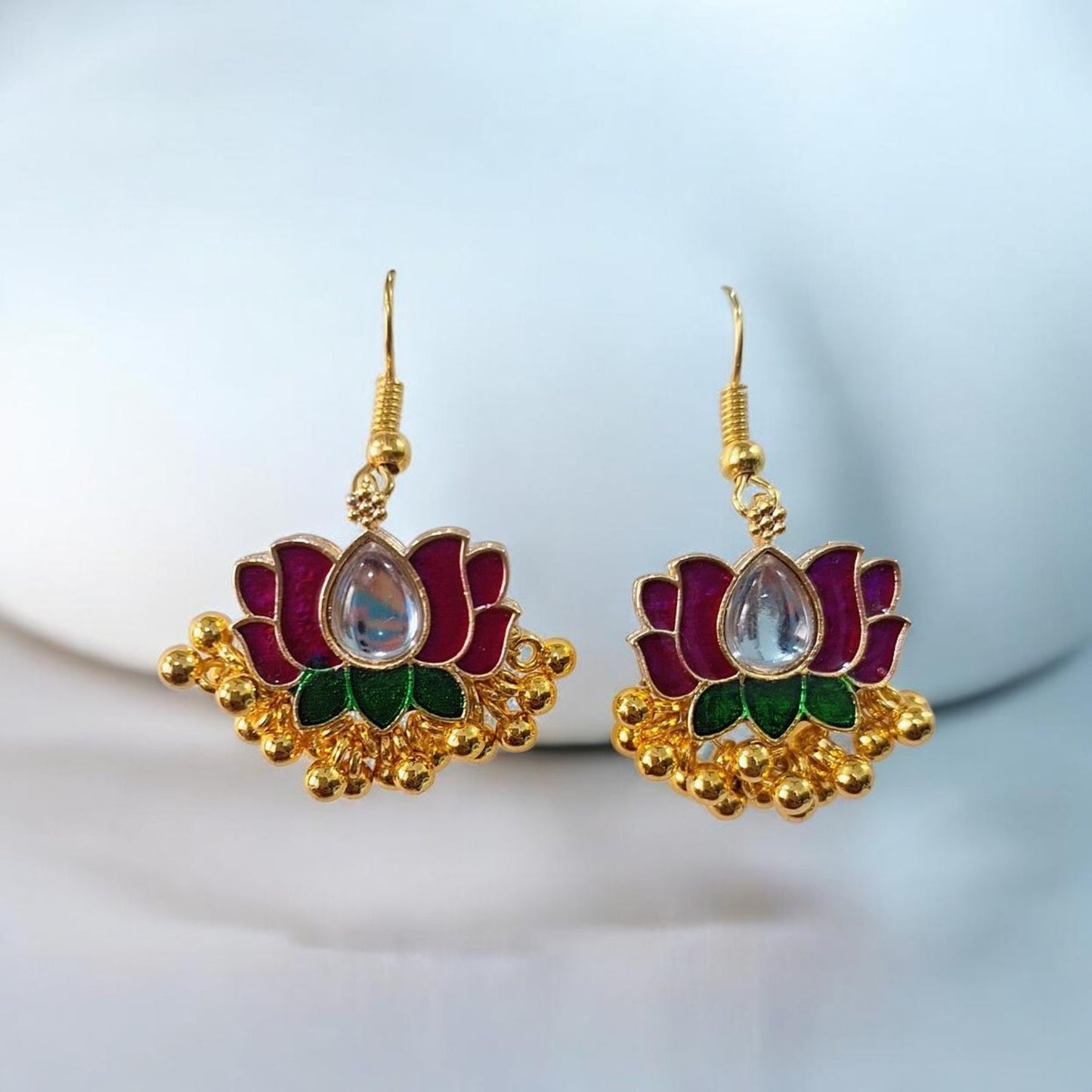 A pair of gold-toned lotus-shaped earrings with magenta-pink petals, green enamel accents, and a clear teardrop-shaped stone in the center, displayed on a white surface.