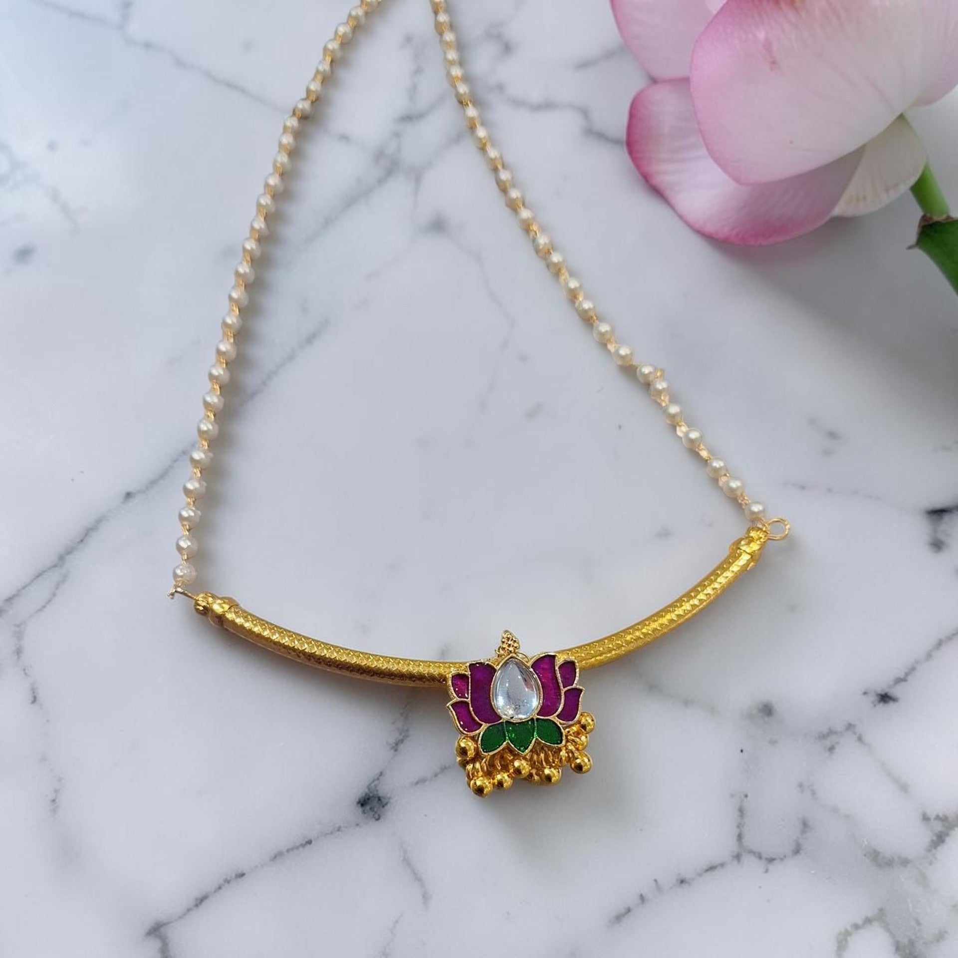 A gold-plated necklace featuring a curved bar and a central lotus pendant with purple and green enamel details and a clear central stone, displayed on a white marble surface with a pink flower in the corner.