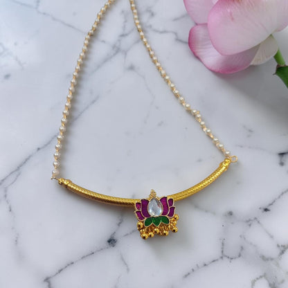 A gold-plated necklace featuring a curved bar and a central lotus pendant with purple and green enamel details and a clear central stone, displayed on a white marble surface with a pink flower in the corner.