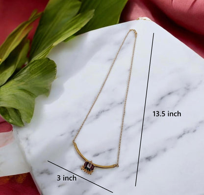 Gold necklace with a curved bar and a small central charm, displayed on a white marble surface with green leaves and pink fabric in the background, with black lines and text showing dimensions of 13.5 inch length and 3 inch width.