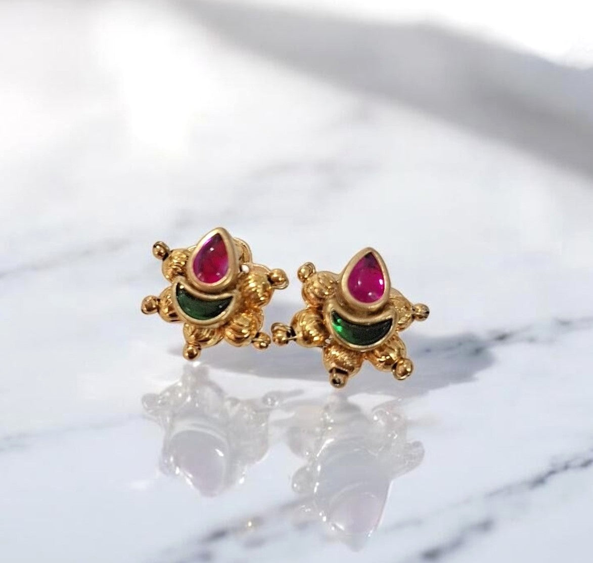 A pair of gold-toned traditional Indian earrings shaped like small decorative figures, featuring teardrop-shaped pink stones and green stone accents, displayed on a white marble surface.