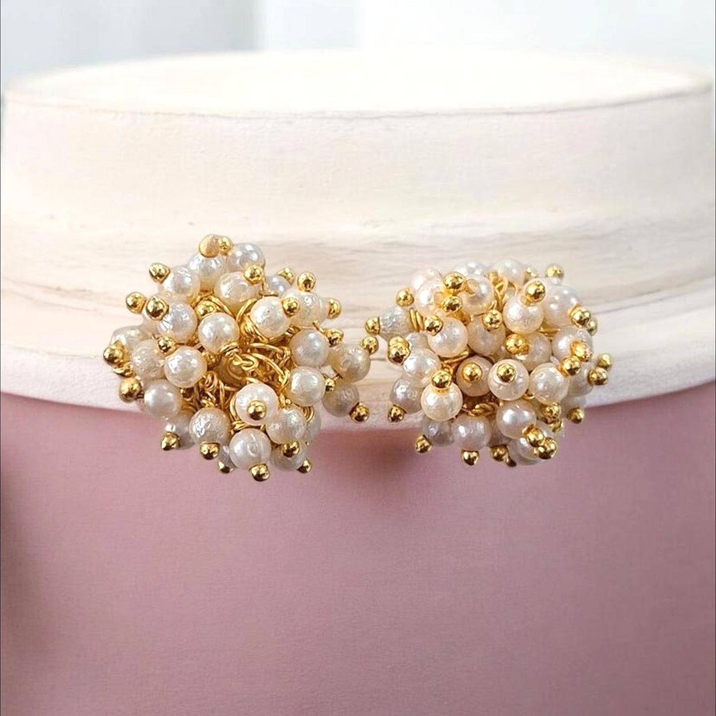A pair of gold and pearl cluster stud earrings displayed on a white ribbon against a soft pink background.