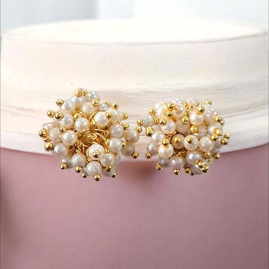 A pair of gold and pearl cluster stud earrings displayed on a white ribbon against a soft pink background.