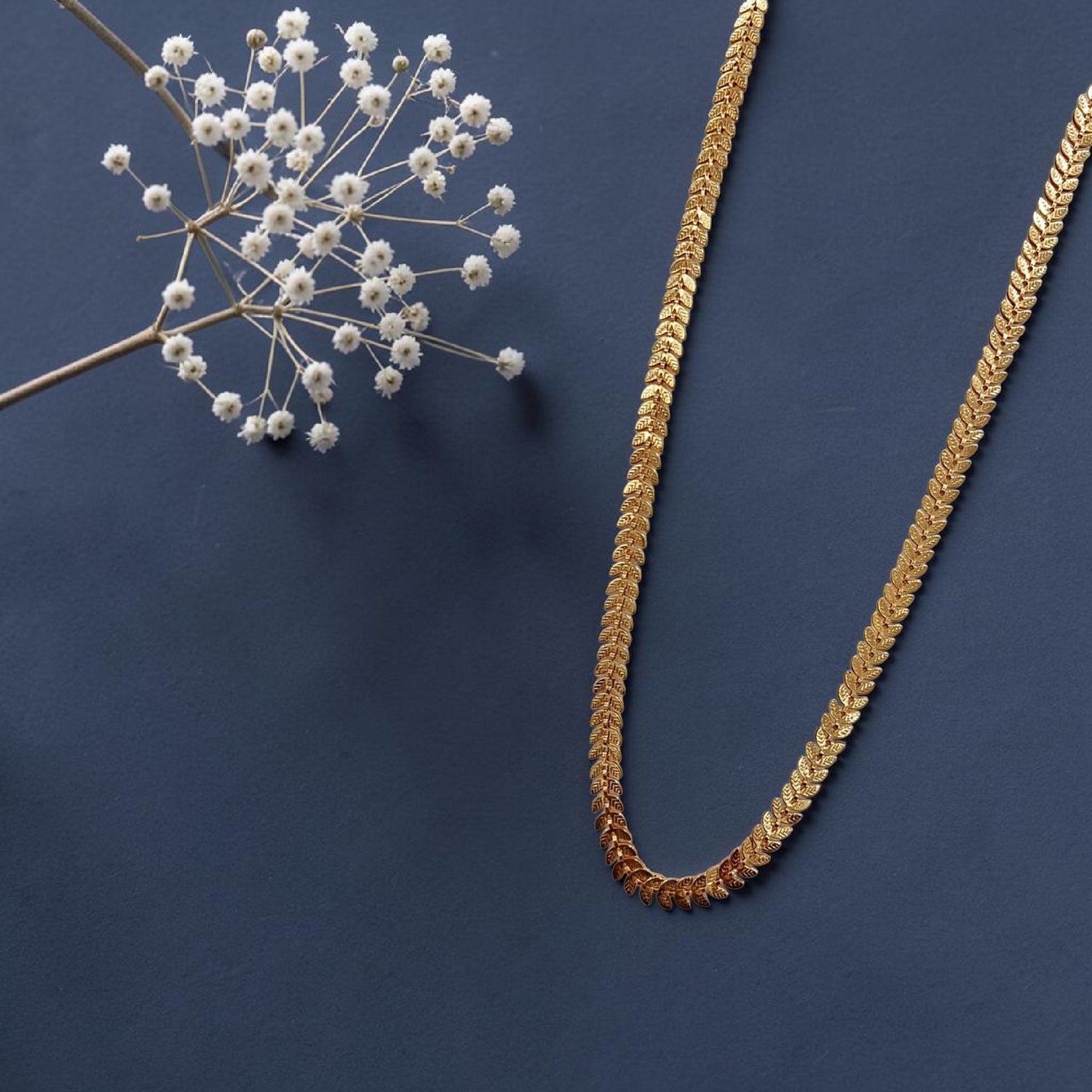 Uparna gold plated leaf chain long necklace laid flat on a navy blue background with dried white wildflowers