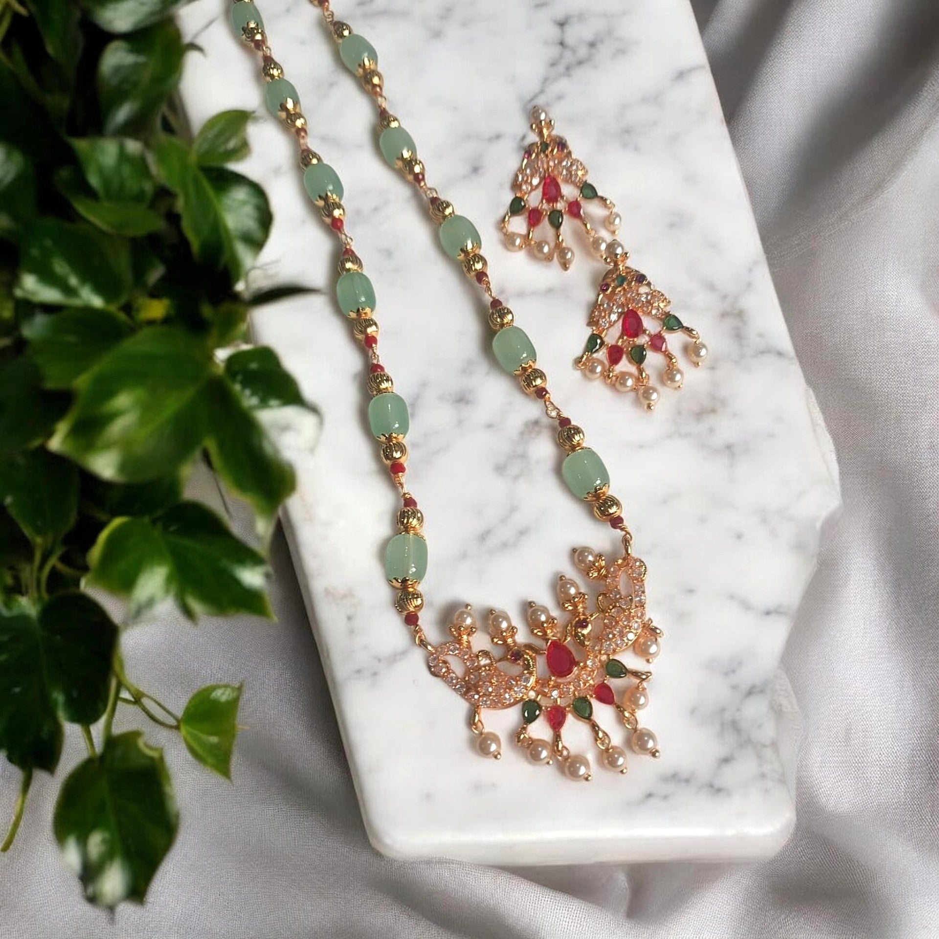 Gold-plated Indian ethnic jewelry set featuring a necklace and matching earrings with light green stone beads, red stones, and small pearl-like drops, displayed on a white marble block next to green leaves.