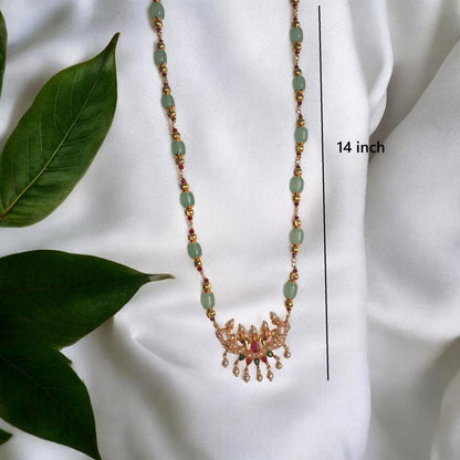 A gold-toned beaded necklace with light green oval beads and a decorative pendant, displayed on a white fabric background with green leaves. A vertical measurement line and text indicate the necklace is 14 inches long.