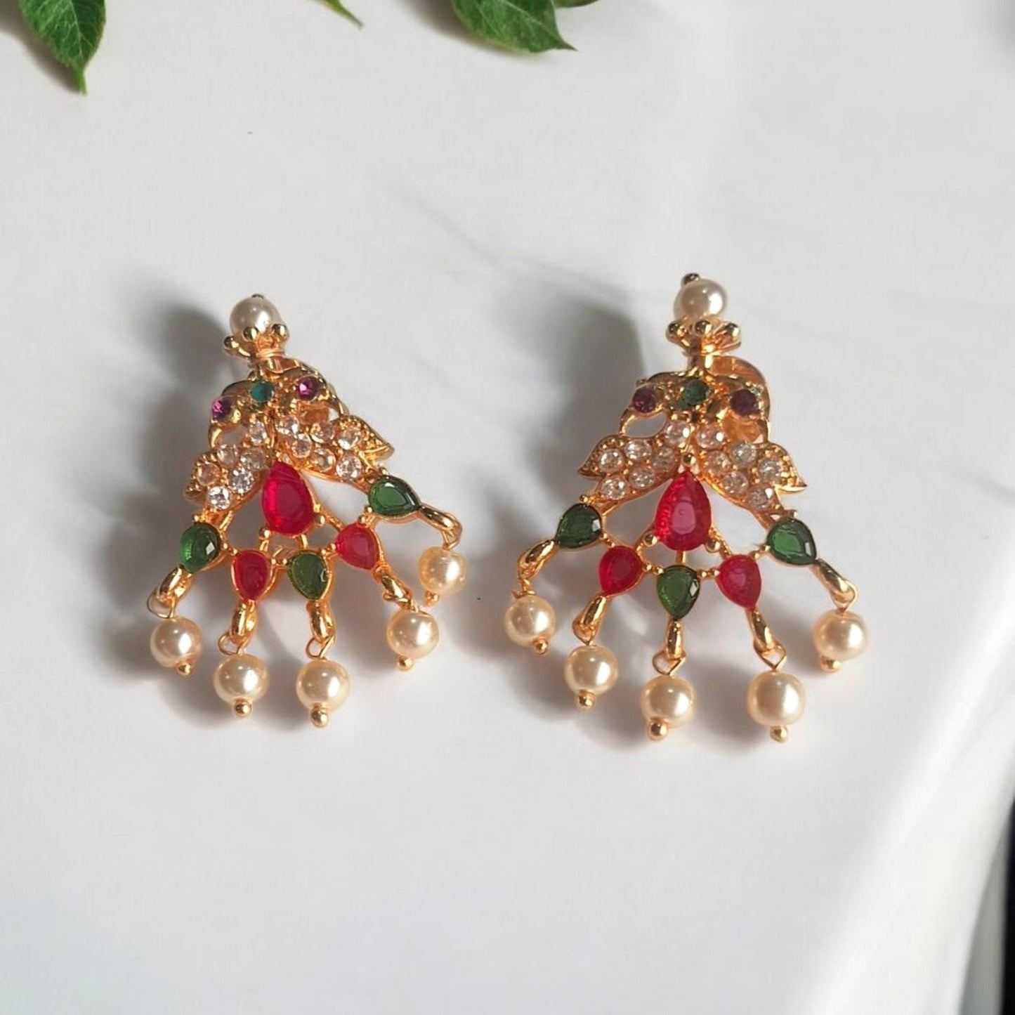 A pair of gold-toned traditional Indian chandbali earrings decorated with red and green stones and white pearl-like drops, displayed on a white marble surface with green leaves in the corner.