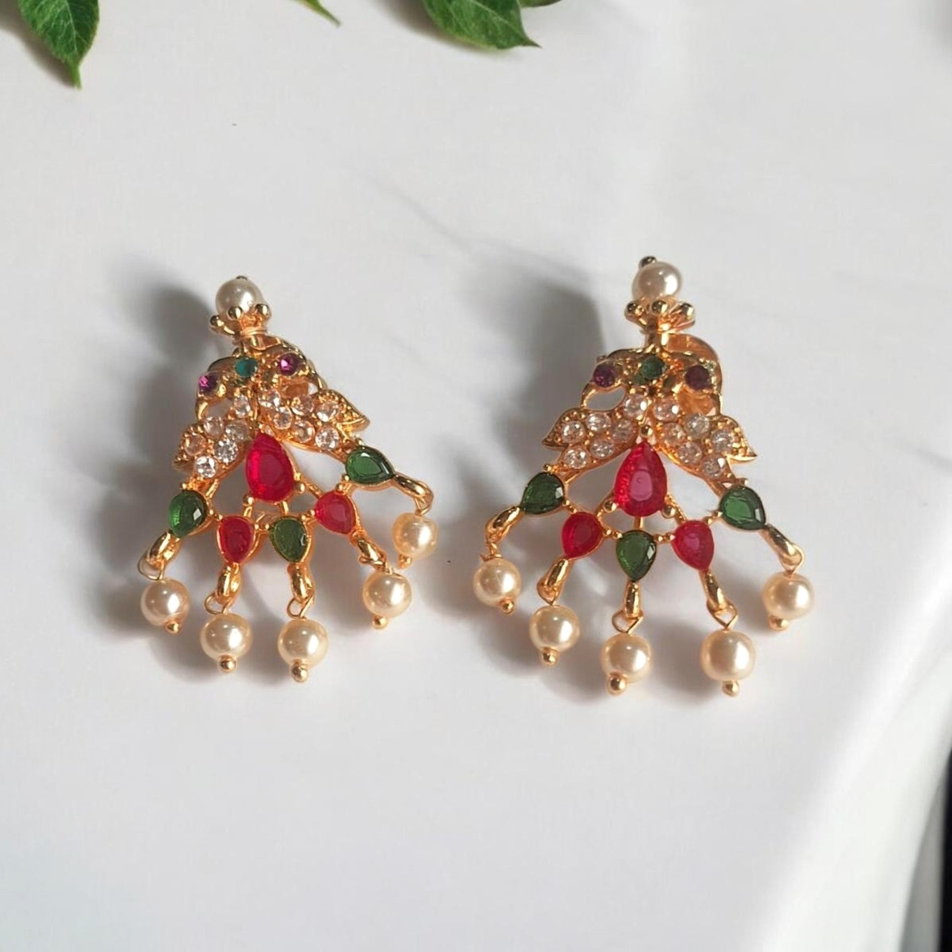 A pair of gold-toned traditional Indian chandbali earrings decorated with red and green stones and white pearl-like drops, displayed on a white marble surface with green leaves in the corner.