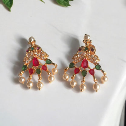 A pair of gold-toned traditional Indian chandbali earrings decorated with red and green stones and white pearl-like drops, displayed on a white marble surface with green leaves in the corner.