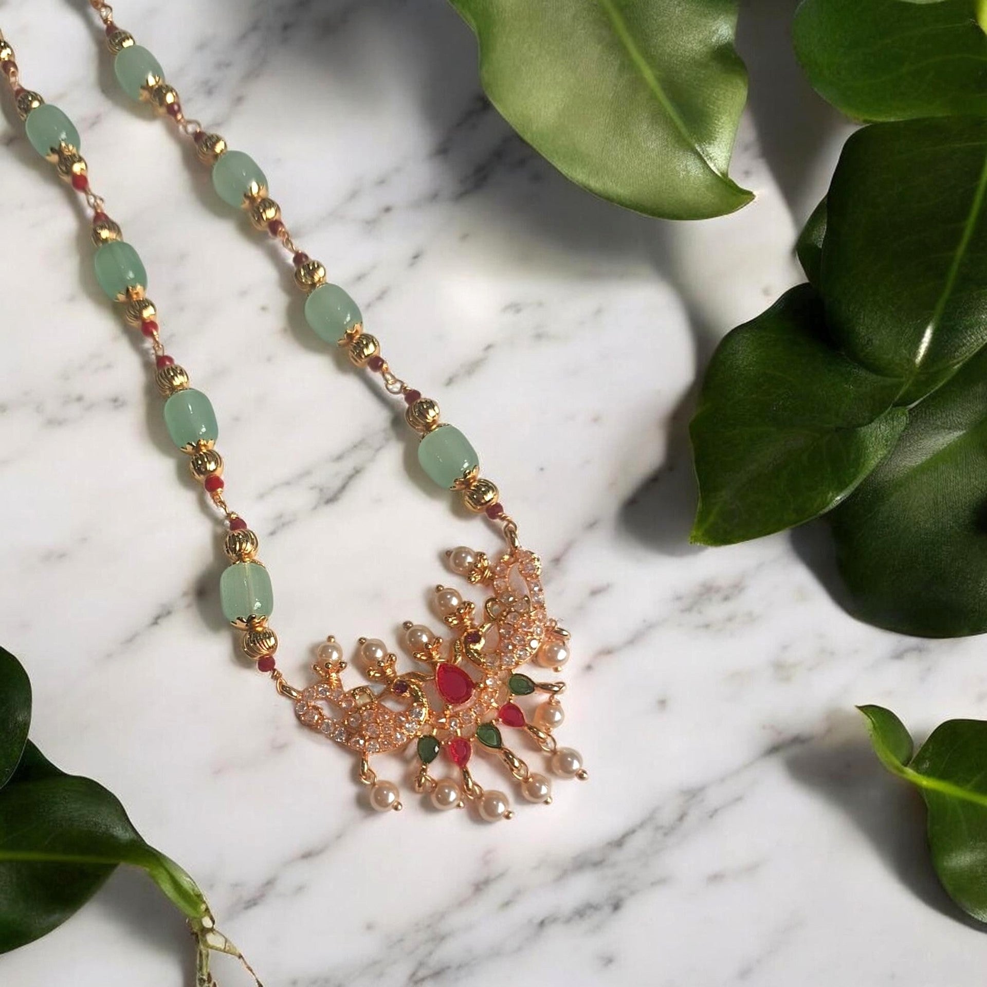 A gold-toned ornate pendant necklace with light green beads, small red and green stones, and pearl-like drops, arranged on a white marble surface with green leaves around it.