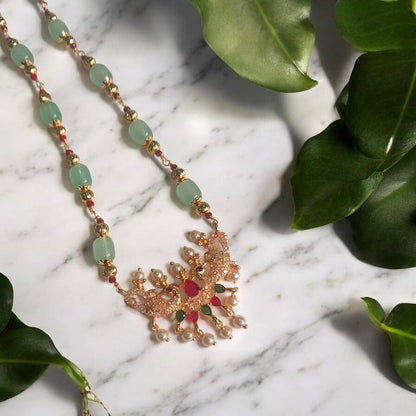 A gold-toned ornate pendant necklace with light green beads, small red and green stones, and pearl-like drops, arranged on a white marble surface with green leaves around it.