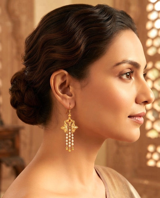 A woman in profile wearing a dangling gold earring with pearl-like bead drops, set against a warm, softly blurred indoor background.
