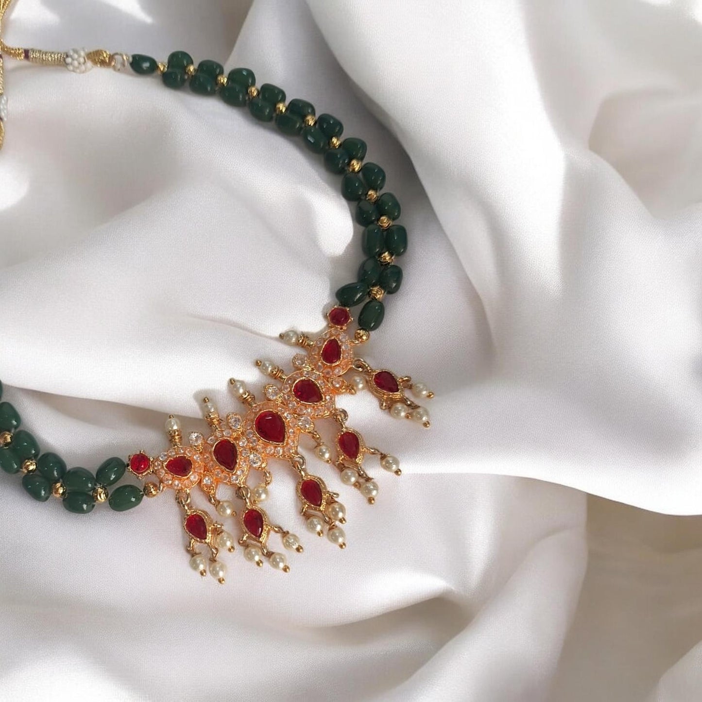 A close-up of an ornate necklace draped on white satin fabric. The necklace features dark green faceted beads, a gold-toned filigree centerpiece with red gemstone accents, and small clear stones.
