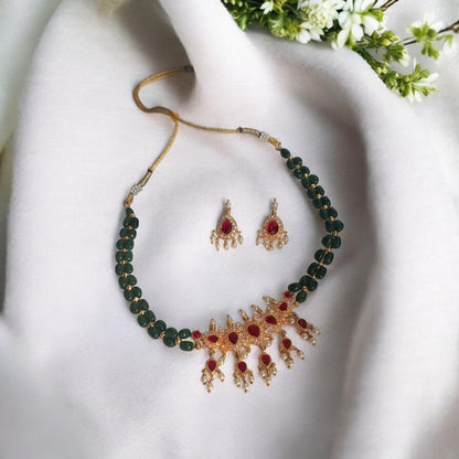 Gold and dark green beaded necklace with a matching pair of dangle earrings, styled on a white fabric background with small white flowers.