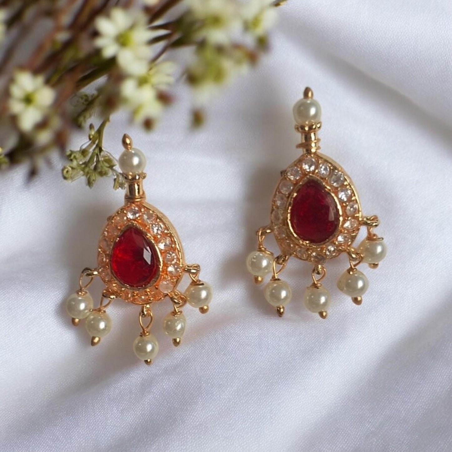 A pair of gold-toned traditional Indian chandelier earrings with red teardrop-shaped gemstones and small white pearl-like beads, displayed on a white fabric background with small white flowers.