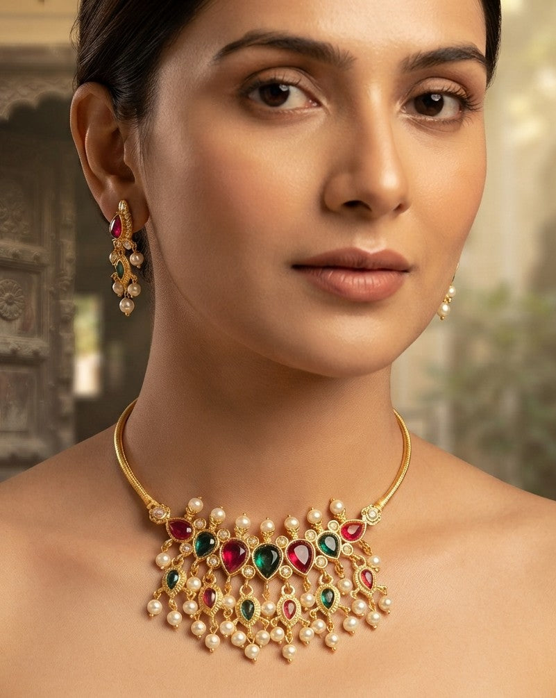 A close-up of a woman wearing a gold-plated statement necklace and matching drop earrings. The necklace features heart-shaped red and green gemstones surrounded by white faux pearls, while the matching earrings feature red stones and pearl clusters on a gold setting.