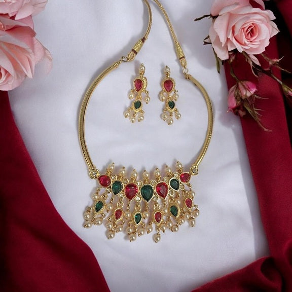 Gold-toned traditional necklace and matching earrings with red and green stone embellishments, arranged on a white fabric background with pink roses and red draped fabric.