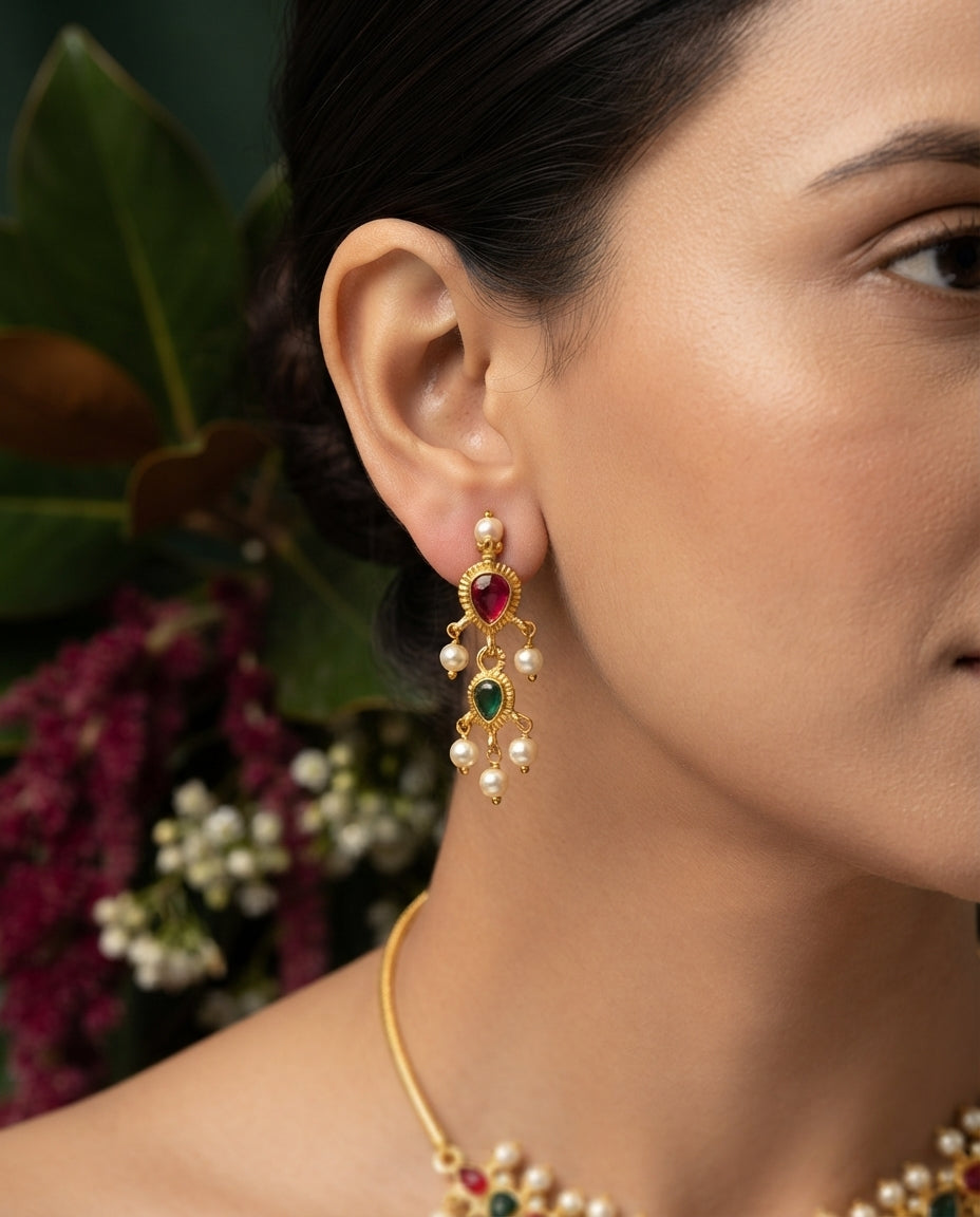 A close-up of a woman wearing a gold-toned traditional Indian chandelier earring adorned with a red ruby stone, green emerald stone, and white pearls, paired with a matching pearl and gemstone necklace.